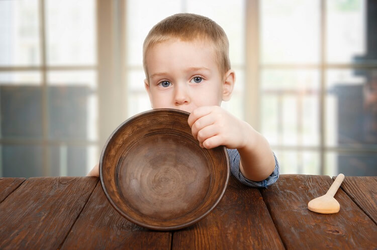 Cute small child boy sitting at wooden table shows empty plate